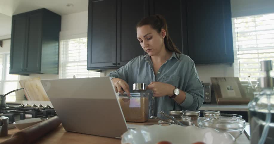A woman adds brown sugar to a measuring cup as she follows a recipe on her laptop