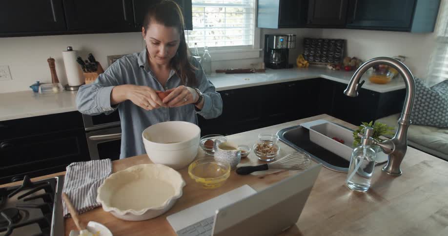 Woman with ponytail and blue shirt cracks egg into white bowl in the kitchen