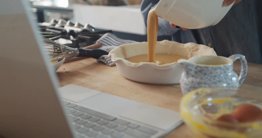 A laptop sits open on a kitchen counter while a woman lifts a bowl and pours filling into a pie plate