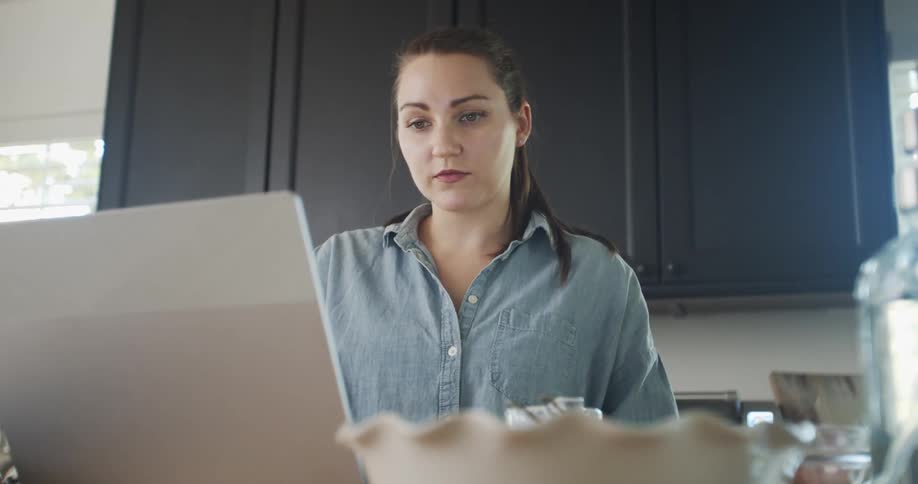 A woman stands in her kitchen at home and looks at her laptop