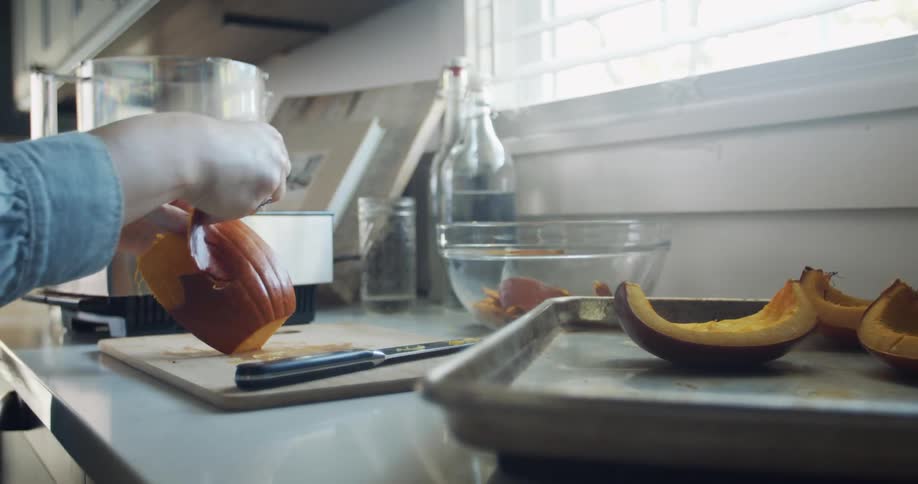 A person peels the skin off a slice of squash on a cutting board on the counter of a home kitchen during the day