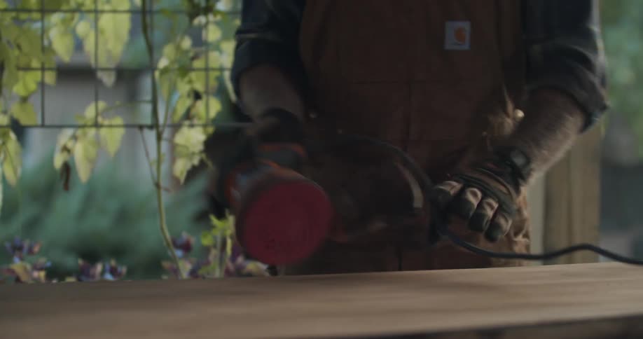 A person with gloves and an electric sander sanding a table with plants the sky and clouds in the background