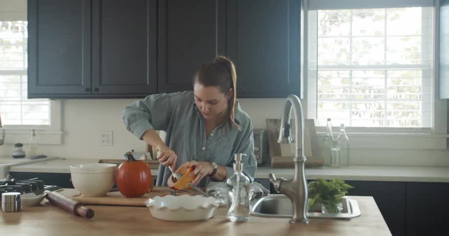 Woman uses a spoon to carve the interior of a pumpkin in the kitchen at home