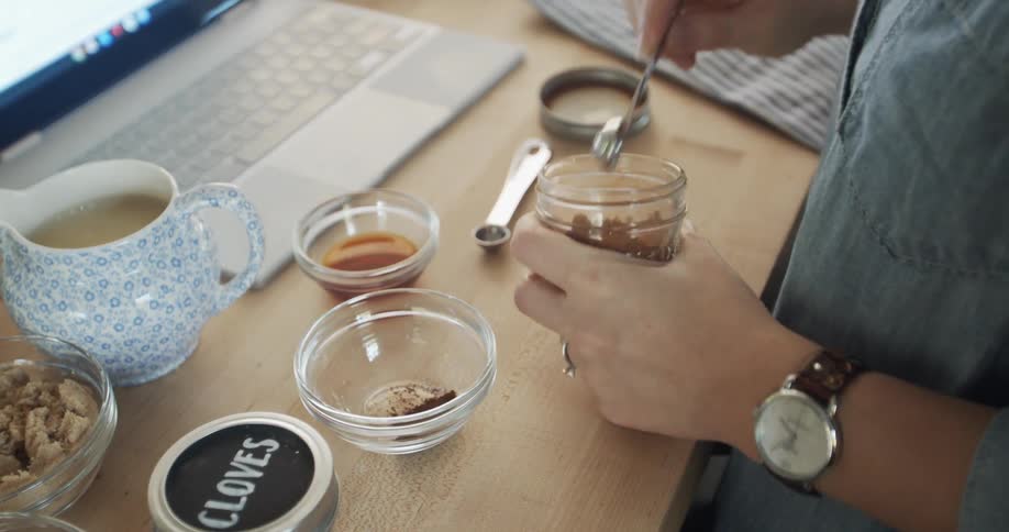 Woman unscrews lid from mason jar full of a spice and then starts to measure with a measuring spoon