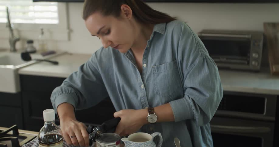 A woman stands at a kitchen counter opening a can with a can opener