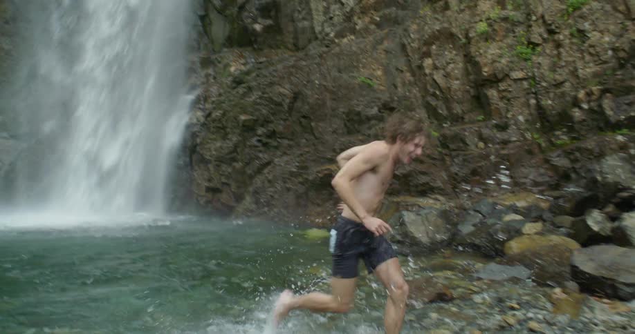Man wearing swim trunks runs out of pool at base of waterfall smiling