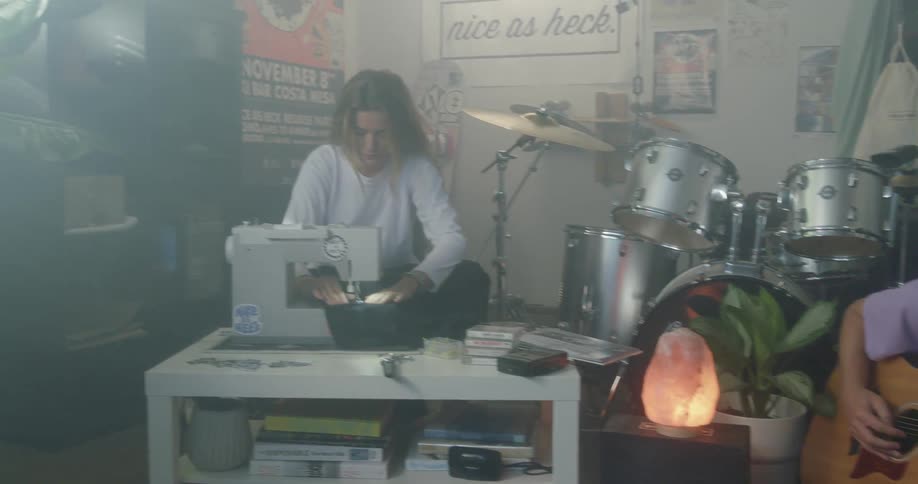 Four people in a house a woman lying on the couch next to a man playing a guitar who sits by a woman sewing and then another man sits at a table reading