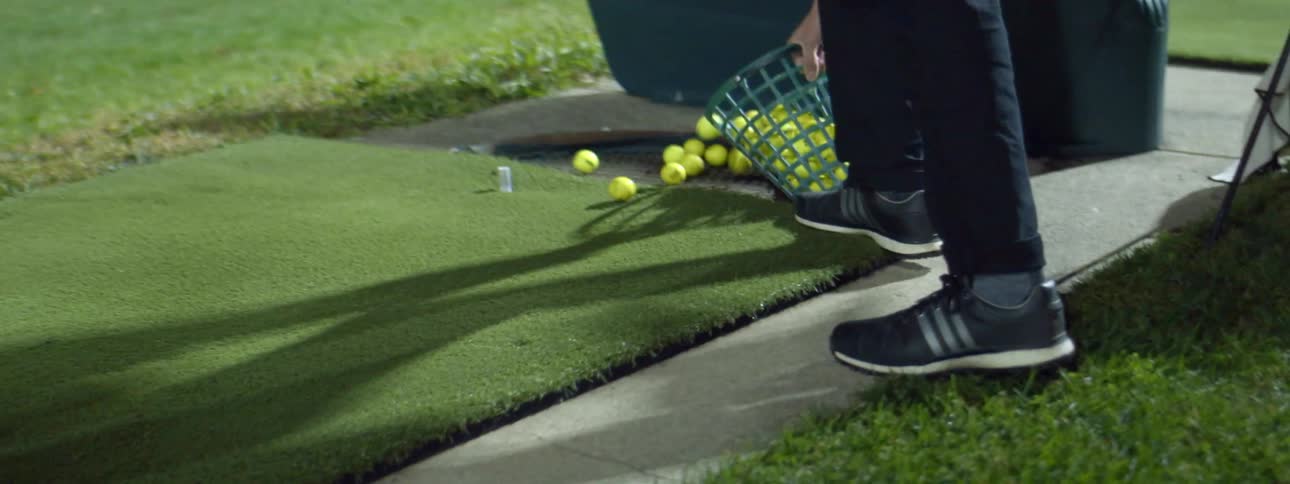 Man empties a bucket of golf balls over the grass in a golf driving range at night