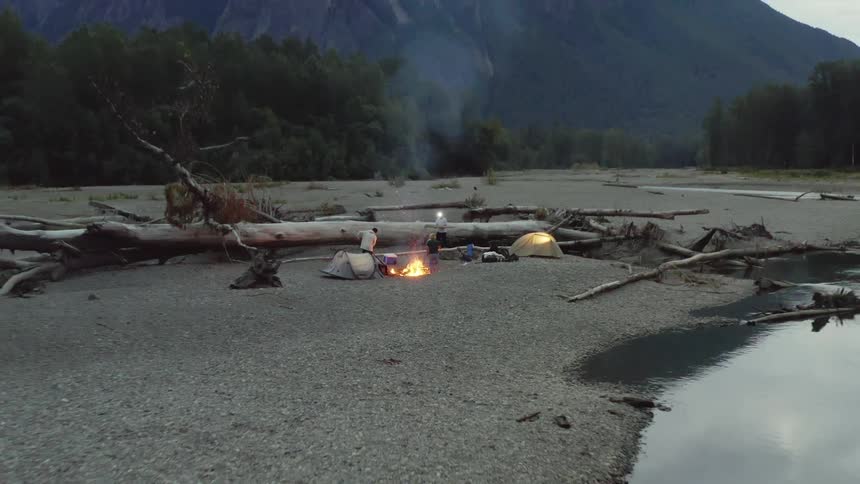 Aerial view of a group of friends camping around a campfire near a river in the pine forests on the mountains at evening