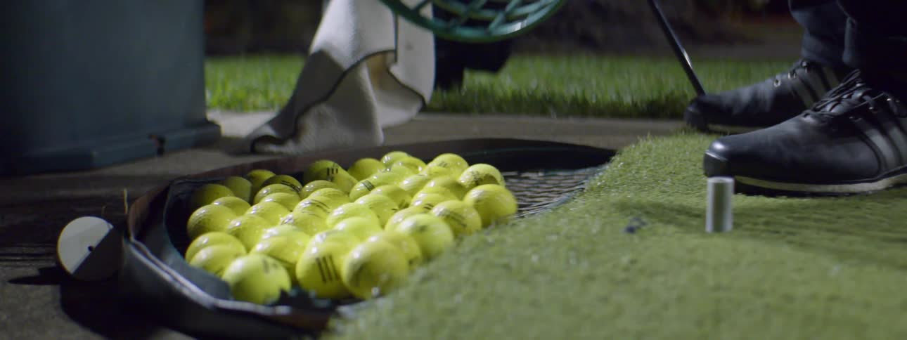 Person pours golf balls out of a basket and onto a rack next to green turf and a golf tee