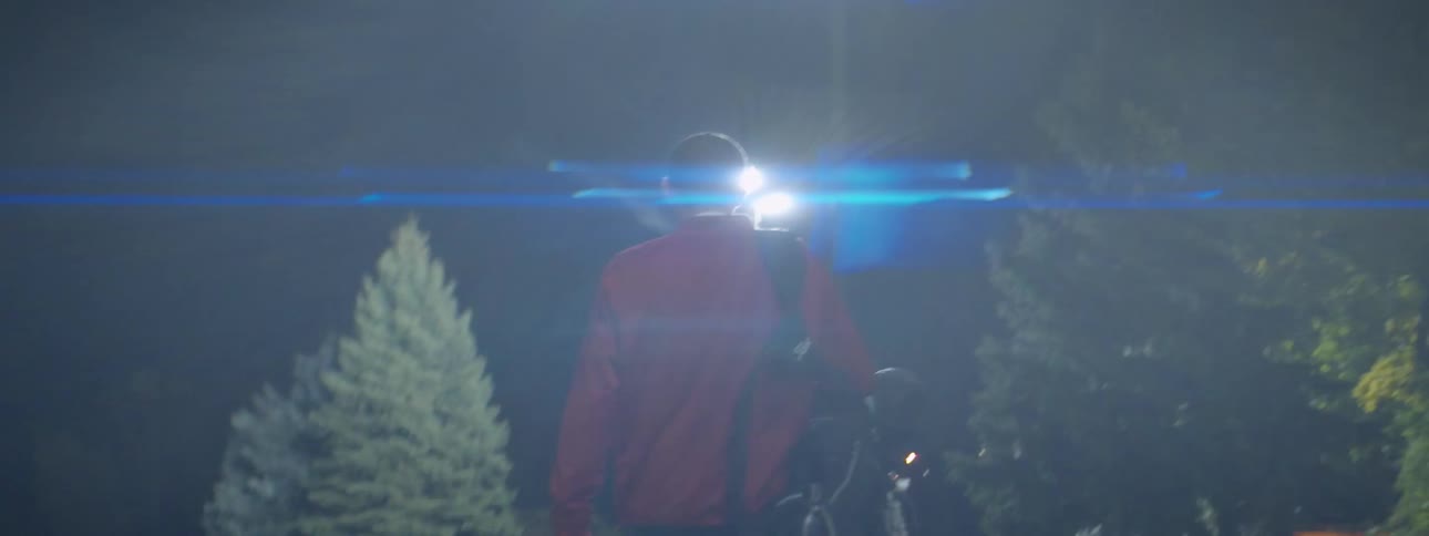 A man walks on a golf course carrying a bag and bucket with golf balls at night