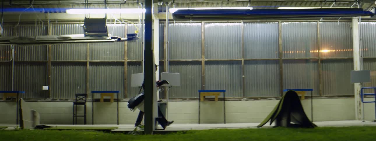 A man walks on a sidewalk past a warehouse carrying a golf bag at night