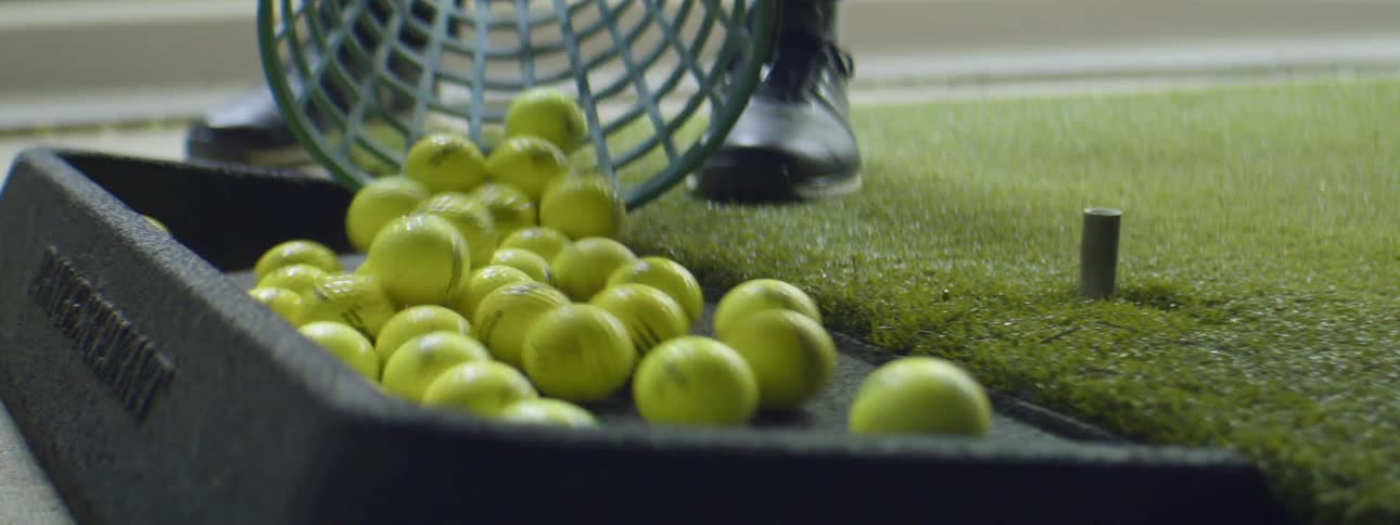 Man empties a bucket full of yellow golf balls inside a golf driving range