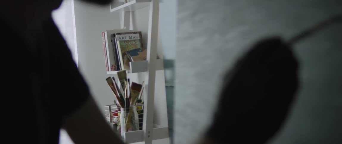 A man paints at an easel and a shelf of art books and tools is shown in the background