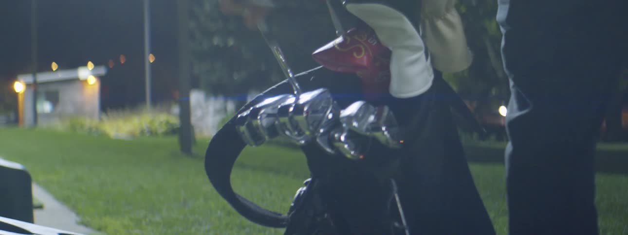 A man standing in a grassy area at night takes a golf club out of a bag while cars drive by in the background