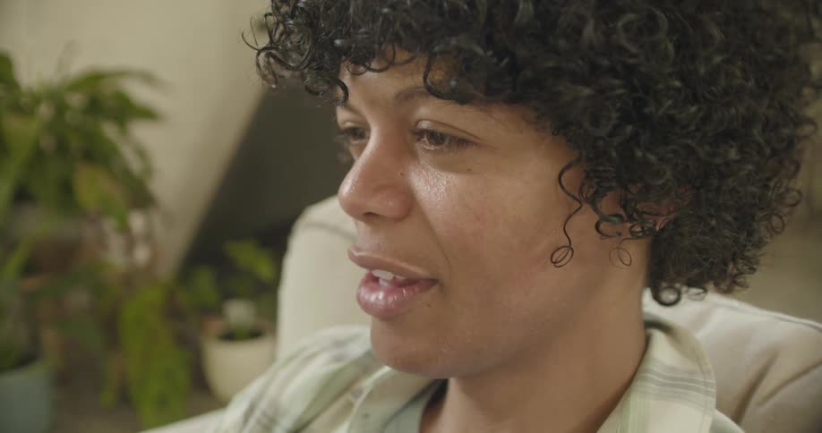 A young man with curly black hair smiles and waves as he sits on a couch during a conversation