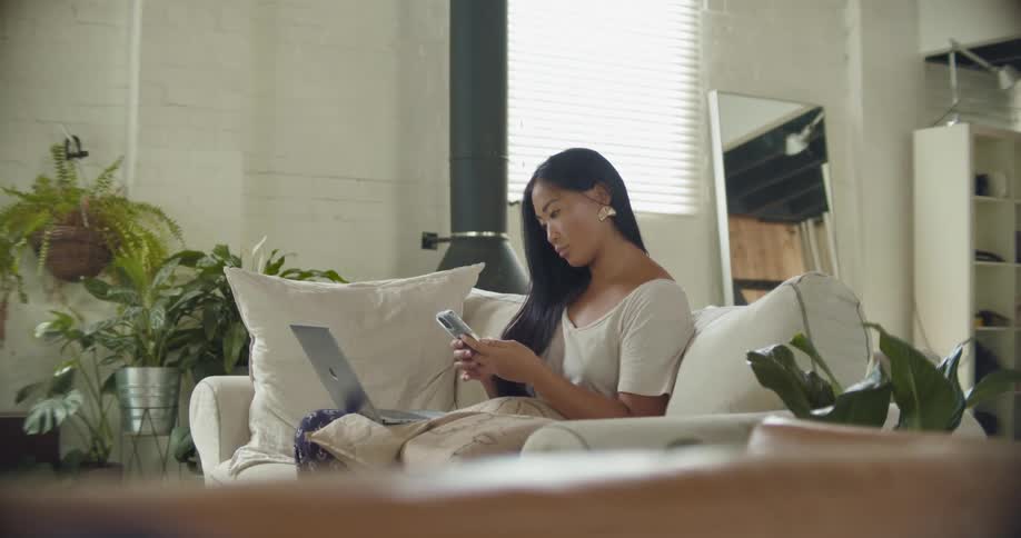 A woman stops typing on her laptop to check her smartphone while sitting on her couch in a bright white living room
