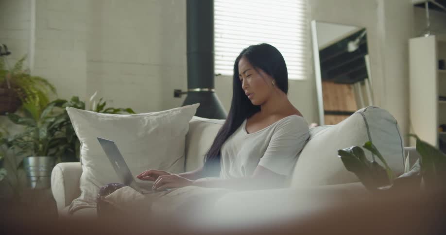 Asian woman with a white shirt sits on the beige sofa and uses a laptop in the living room at home