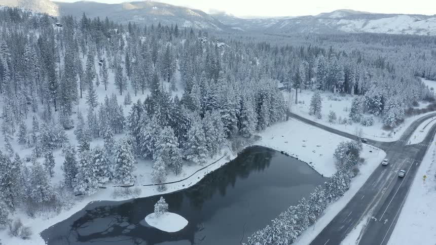 Cars driving on a road near a lake on the snow covered pine tree forest close to the mountains at evening