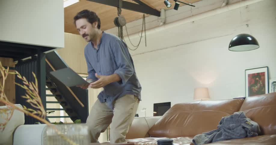 A man walks in to his living room carrying his laptop and waves at the screen as he video chats