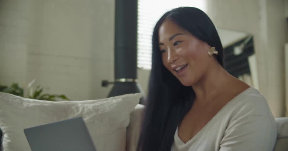 Woman with long hair and white shirt makes video call on laptop in the living room at home