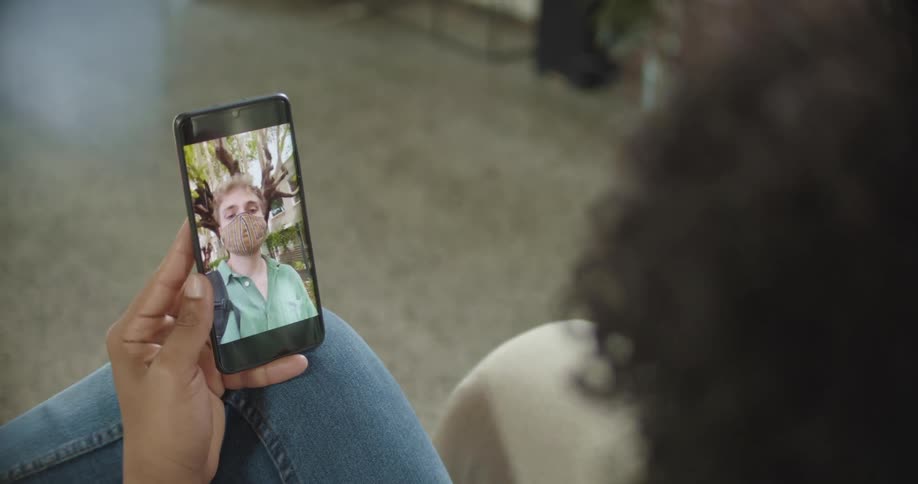Woman with jeans sits on sofa and makes video call on the smartphone with man wearing green shirt and mask