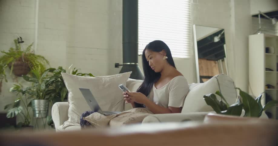 Woman with long hair and white shirt sits on beige sofa uses smartphone and laptop in living room at home