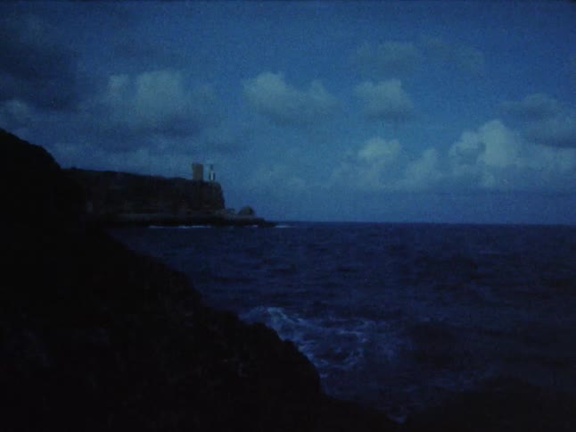Waves rolling in to rocks on the shore and a light flashing in a lighthouse in the distance