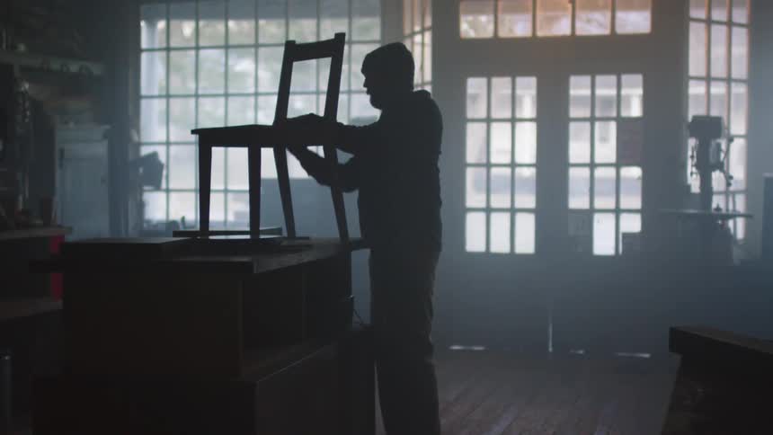 The silhouette of a man using a cloth to polish a wooden chair in a dimly lit workshop during the day