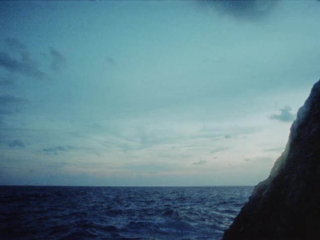 View of the ocean and the horizon at dawn or dusk with some clouds in the sky and a large rock nearby
