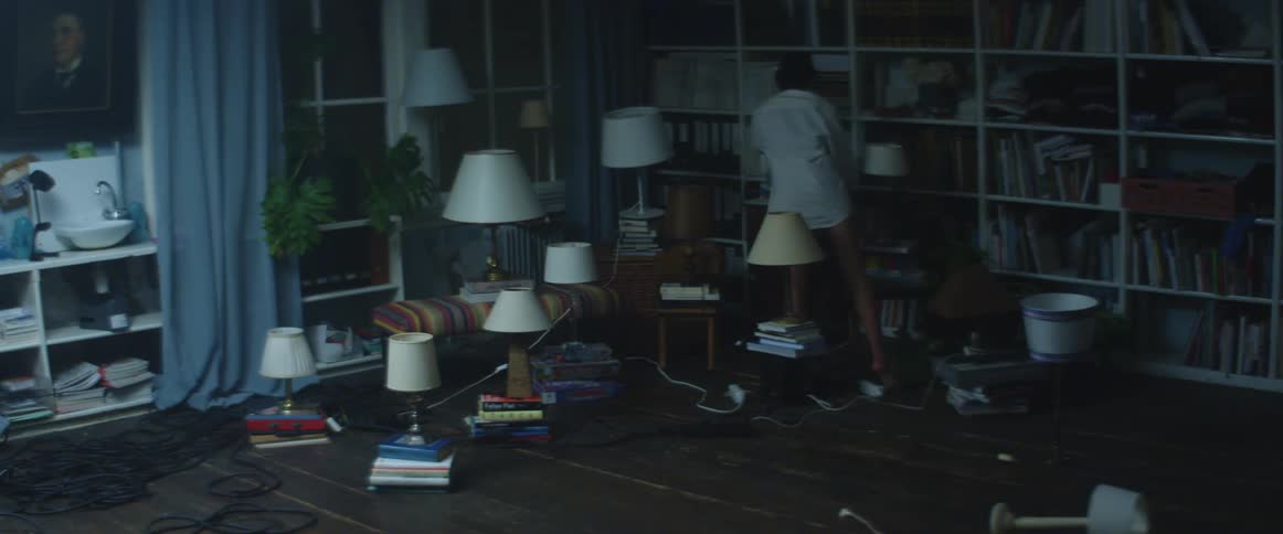 A barefoot pantless woman in a long shirt sets up a lamp in a formation of lamps in a dimly lit room in a house at night