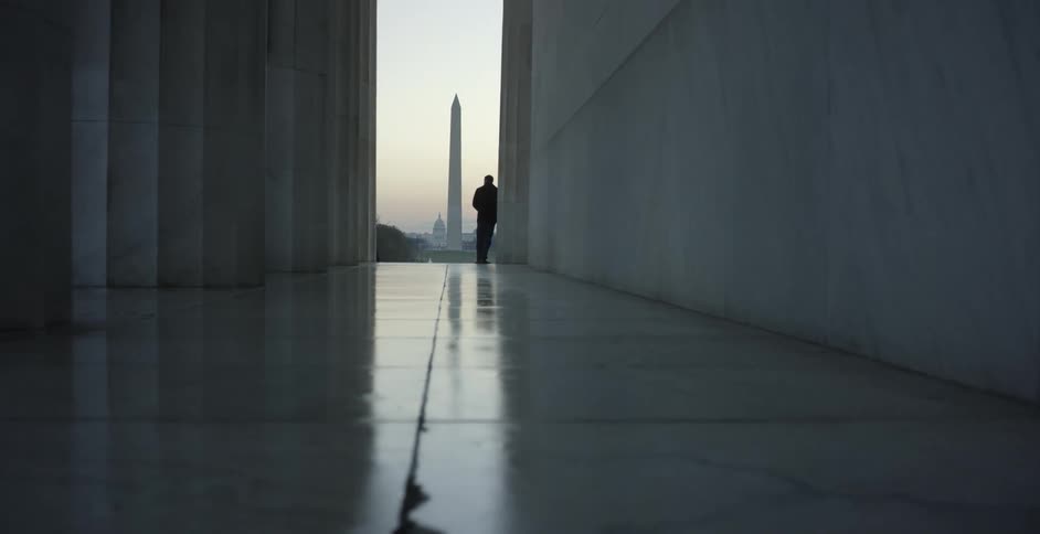 A man stands at the end of a long corridor and looks at a monument