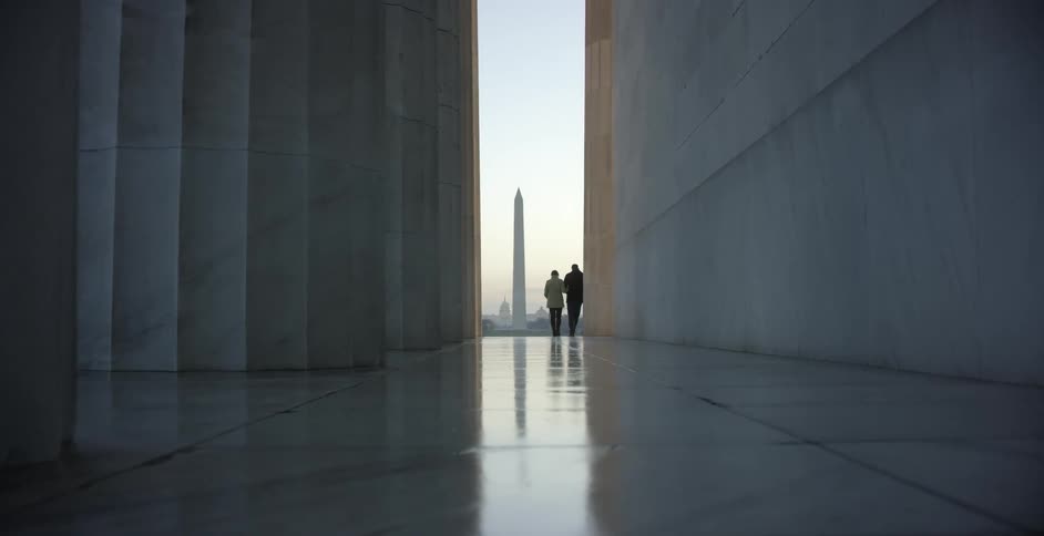 A man and woman hold hands why standing in front of a government building and looking out towards a distant monument in the evening
