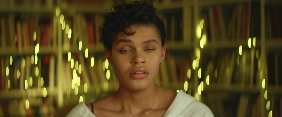 A woman breathes deeply and closes her eyes while sitting in a room filled with shelves and books