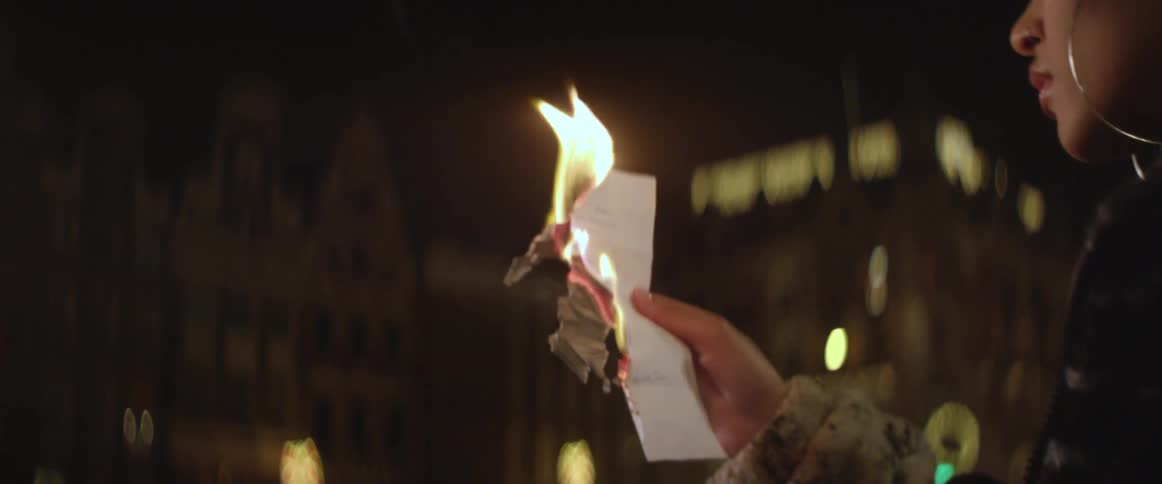 A woman holds a burning sheet of paper while standing outdoors in a metropolis at night