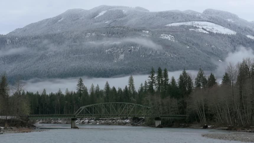 Bridge over river with pine trees and snow covered mountains on a foggy day