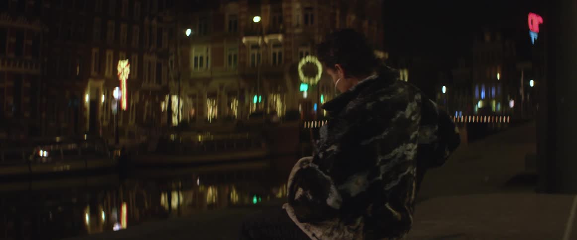 A woman sits on a bench overlooking a town and water at night