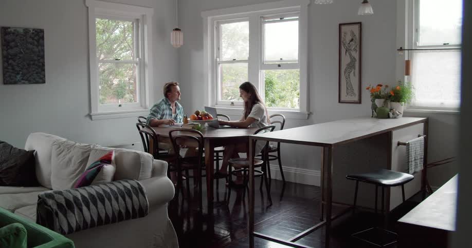 A man and woman speak while sitting at a dining table in a house with a laptop and notebook during the day