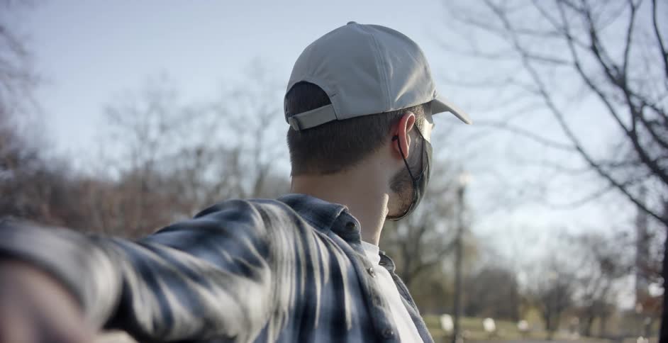 A man stands in a public park with a mask on and waves at another man as he jogs by
