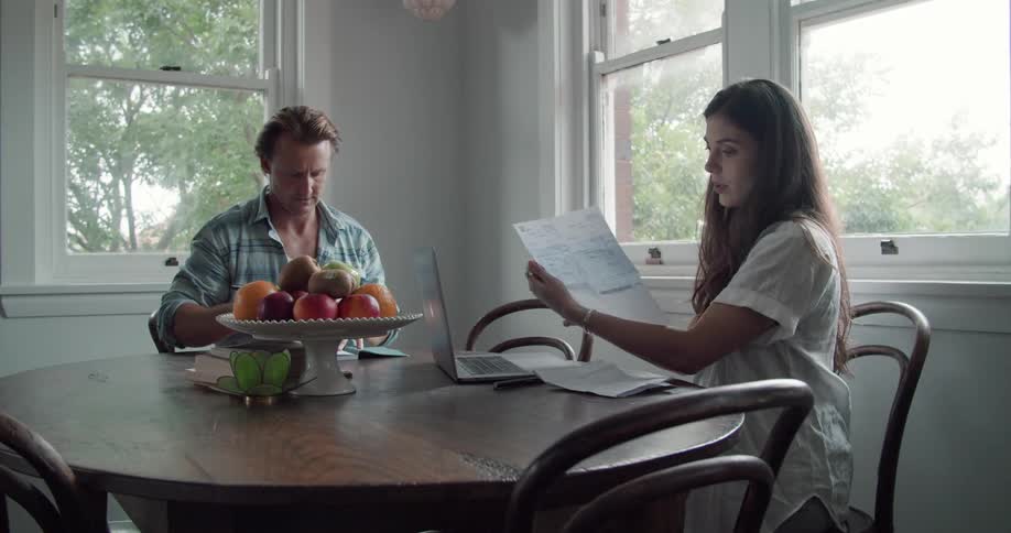 A woman sits at a table at home and types on her laptop and then a man hands her some papers
