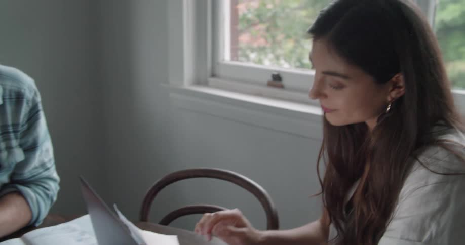 A man and woman sit at a table in a home and he passes her a piece of paper and laughs and she looks at it and a laptop