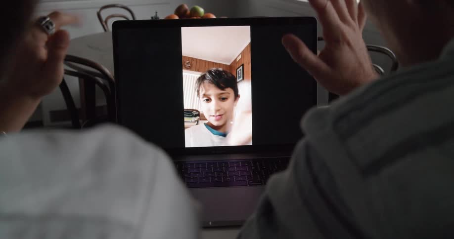 A man and woman sitting in a house during the day use a laptop to wave at a boy through video chat