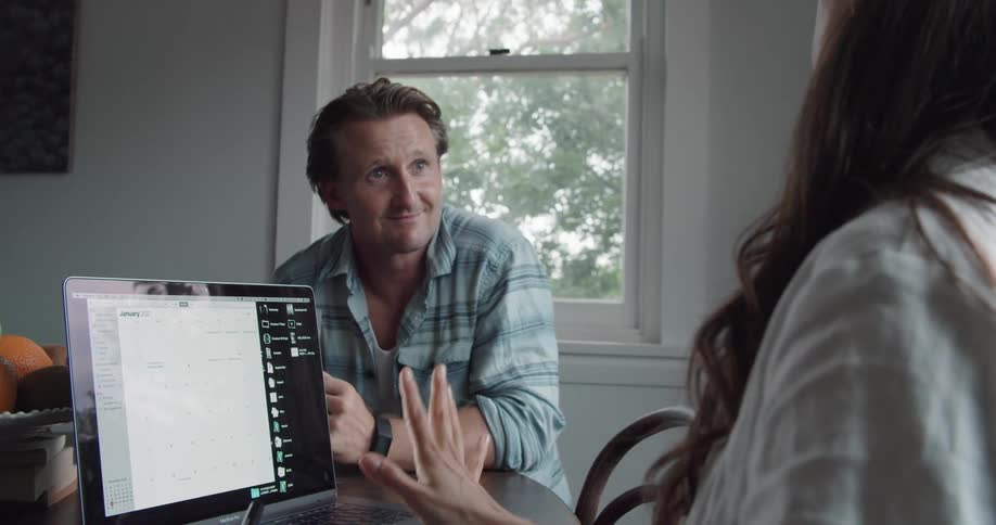 Woman with long hair and white shirt writes in notebook and talks with man wearing turquoise shirt and black wristwatch in the kitchen