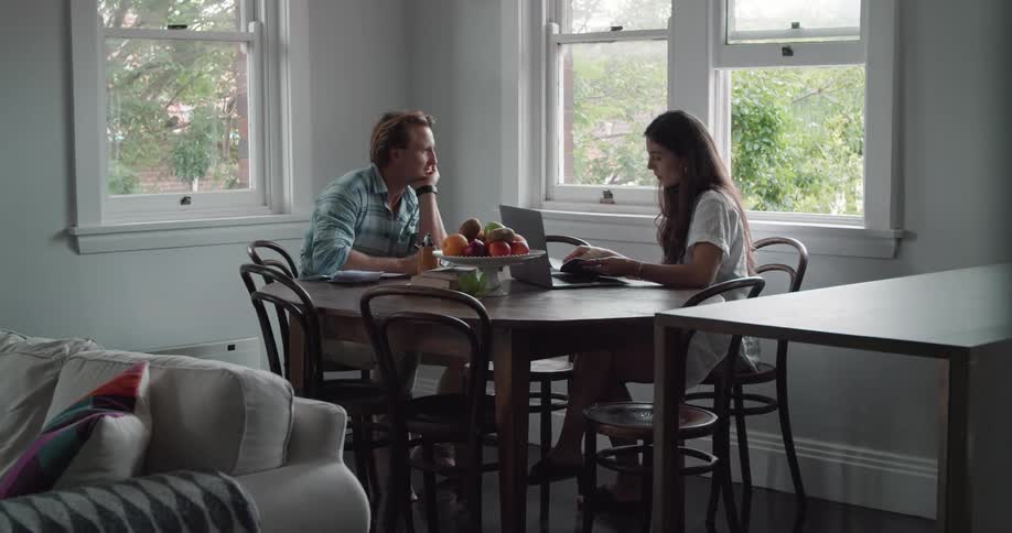 A couple sits at a dining room table next to windows at home working together and taking notes with a laptop at daytime