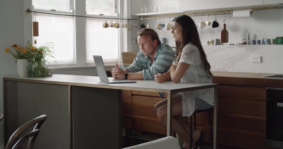 A man puts his arm around a woman and gives her a squeeze as they laugh together and look at a laptop in a kitchen