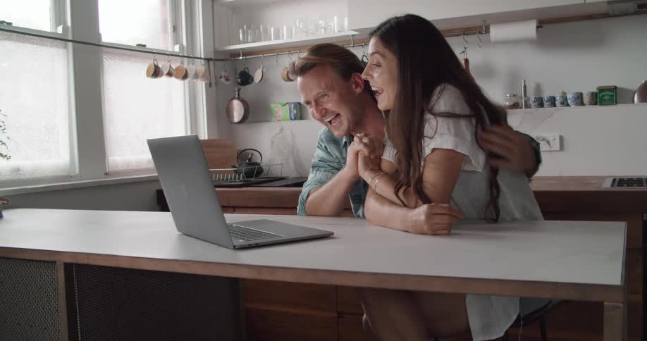 A man and a woman holding hands sitting at a counter in a kitchen video chatting on a laptop computer