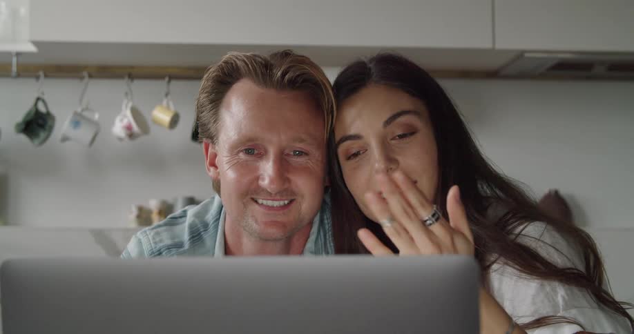 A man and woman leaning against one another kisses to the camera of a laptop sitting before them