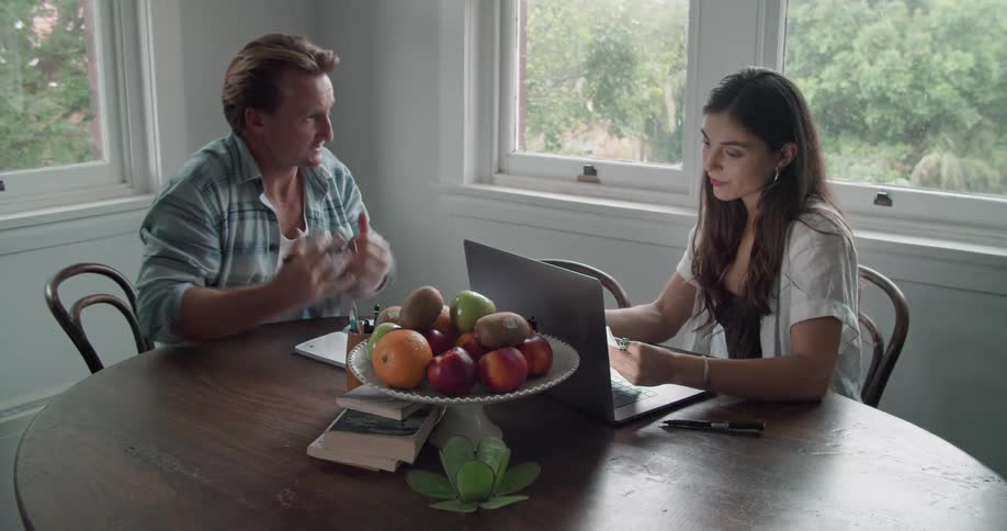 A man and woman sit at a kitchen table with a laptop and a plate of fruit discussing a paper