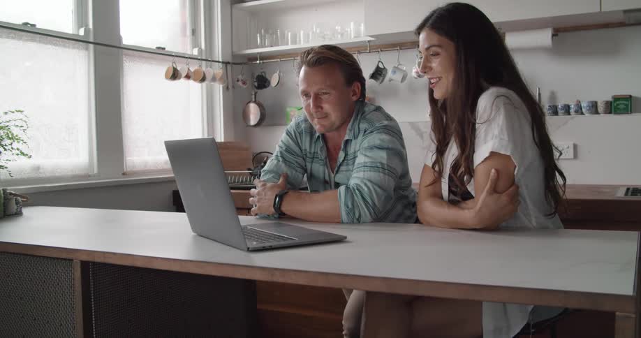 Young couple talking and laughing on a video call in a laptop on the kitchen at home