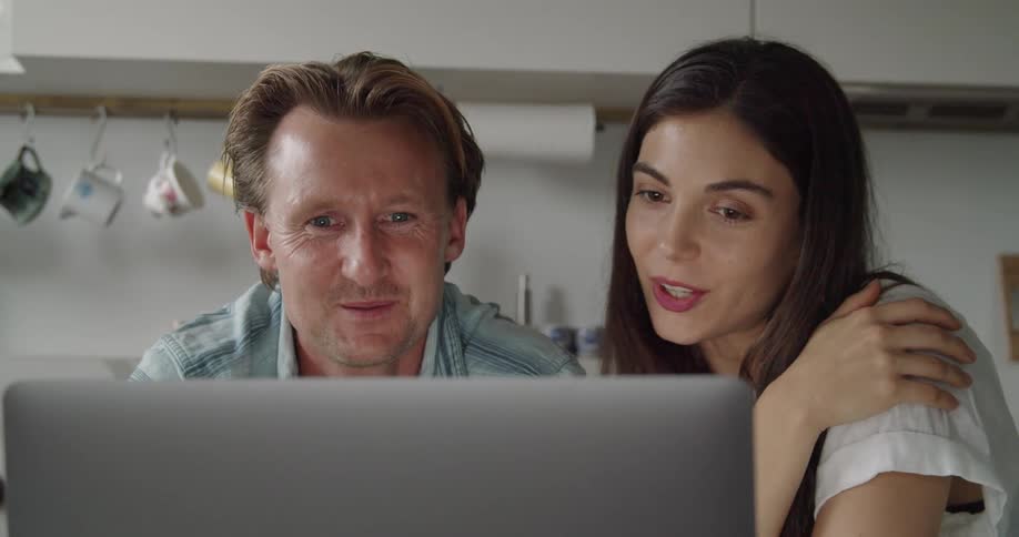 Young couple waving and smiling on a video call in a laptop on the kitchen at home